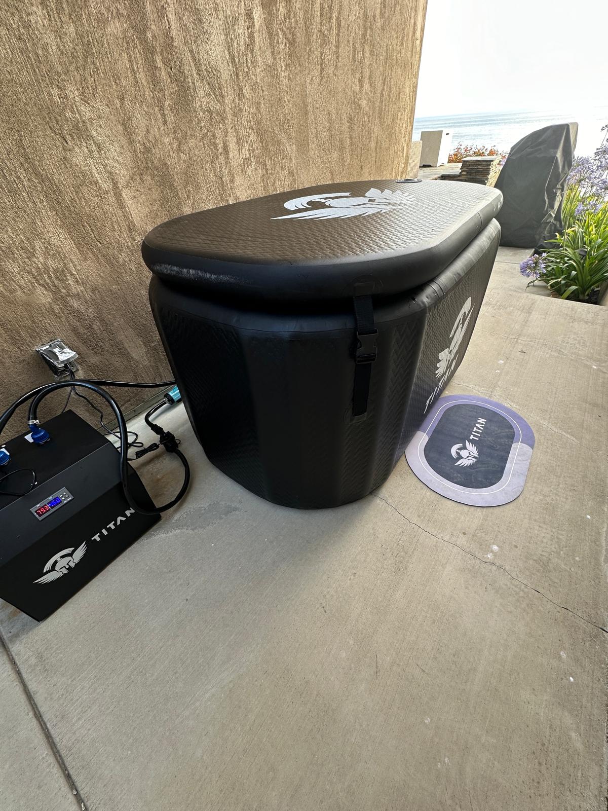 “Triumph XL cold plunge in black with lid closed on an outdoor patio, connected to a Titan chiller on the left; anti-slip mat in front and ocean view in the background.”
