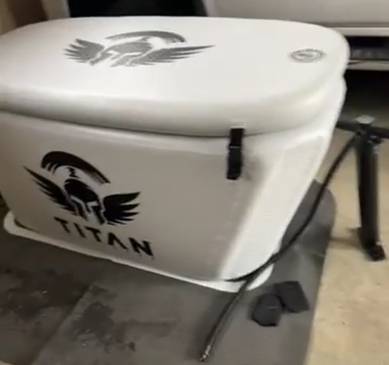 “White Triumph XL cold plunge tub with lid closed and side buckle, featuring the Titan spartan-wing logo; hose resting on an anti-slip mat in an indoor/garage setting.”