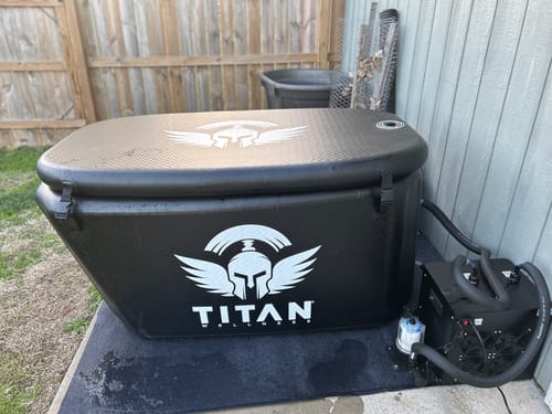 “Triumph XL cold plunge in black with lid closed, connected to a Titan chiller (hoses and filter visible), sitting on a mat beside a house wall and wooden fence in a backyard.”